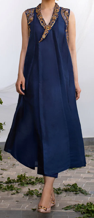 A woman wearing a navy blue dress with gold embroidery and a high neckline, standing on a brick floor with green leaves scattered around her.