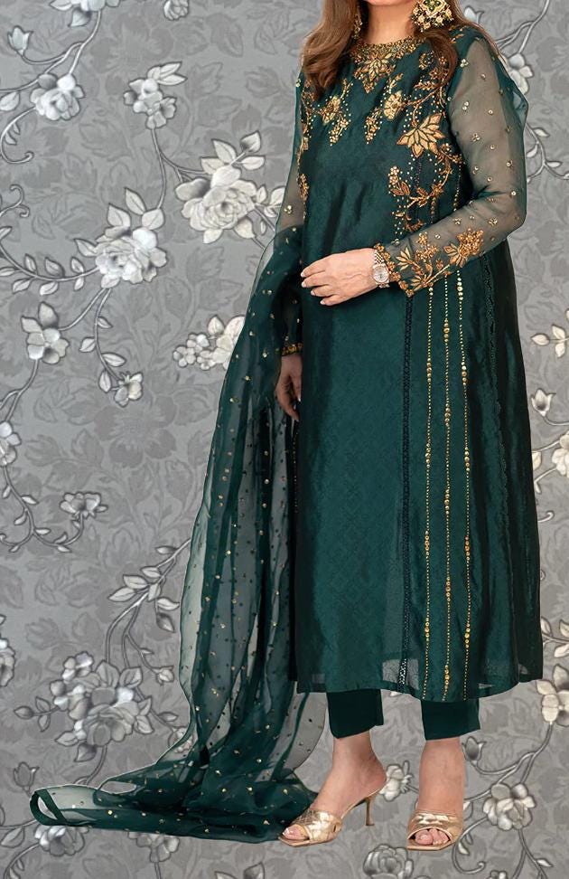 A woman wearing a green and gold dress with intricate embroidery and a matching green shawl, standing against a gray floral background.