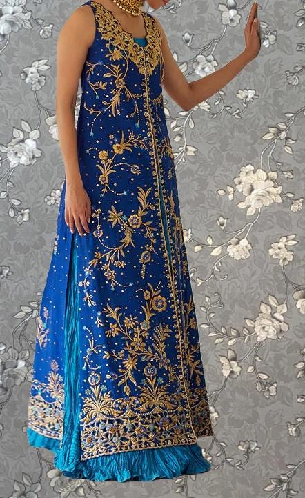 A woman wearing a long, blue dress with gold embroidery stands against a gray floral background.