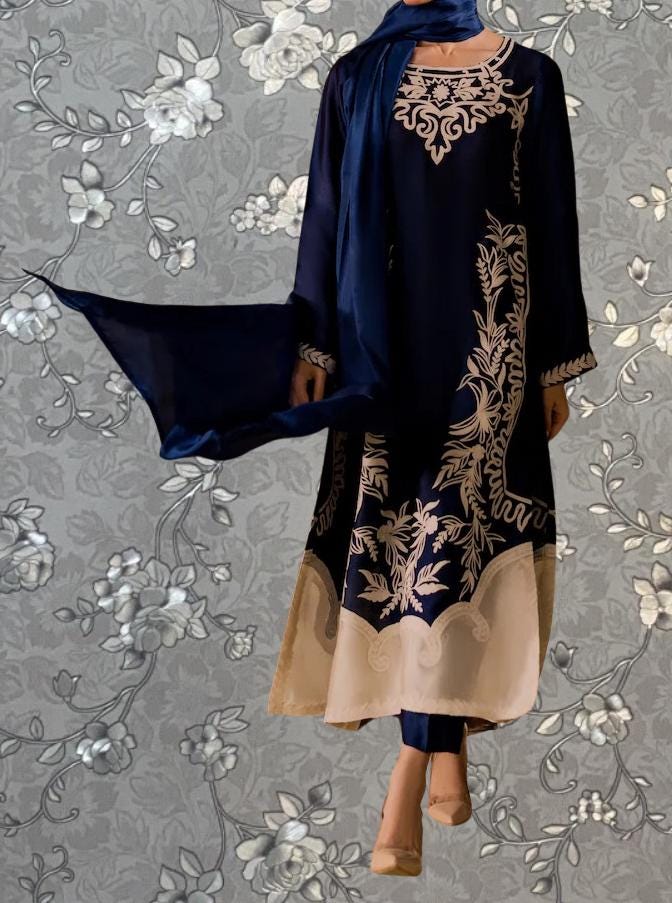 A woman wearing a blue and gold dress with intricate embroidery stands against a gray floral background.