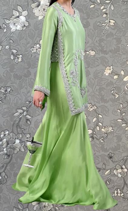 A woman wearing a long, green dress with intricate embroidery and a matching green purse stands against a gray floral background.