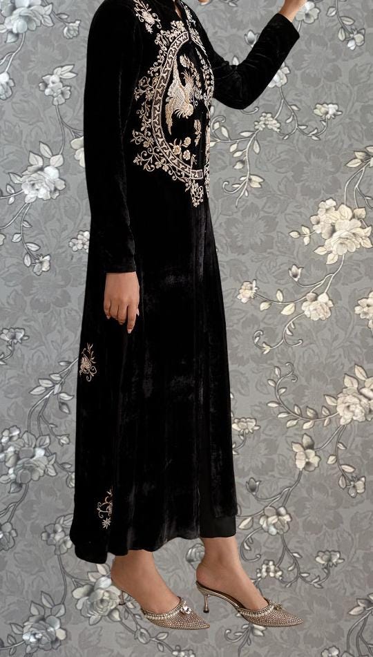 A person wearing a long black dress with gold embroidery stands against a gray floral background.