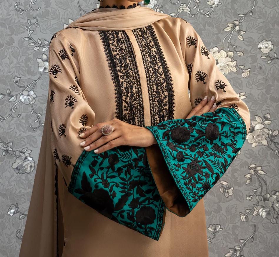 A person wearing a beige dress with black floral embroidery and teal accents, standing against a gray floral background.