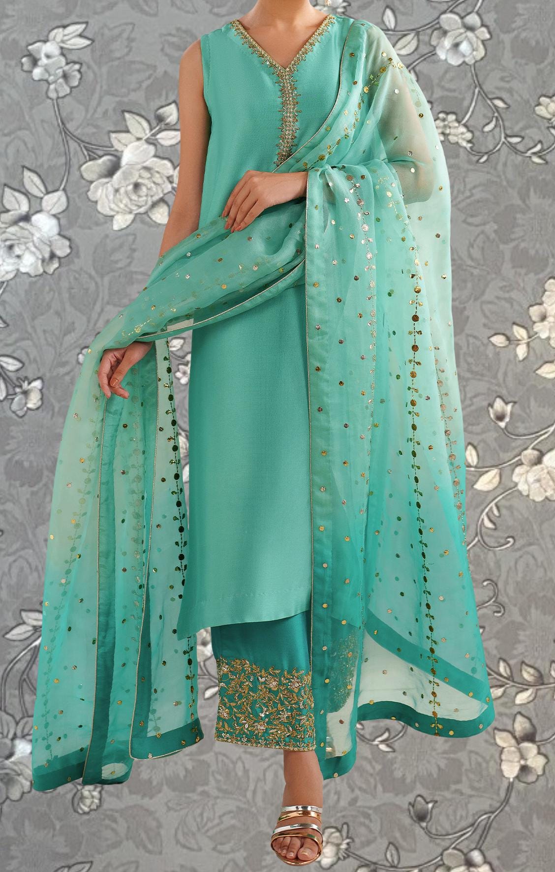 A woman wearing a turquoise dress with a matching turquoise shawl and gold embellishments, standing against a gray floral background.