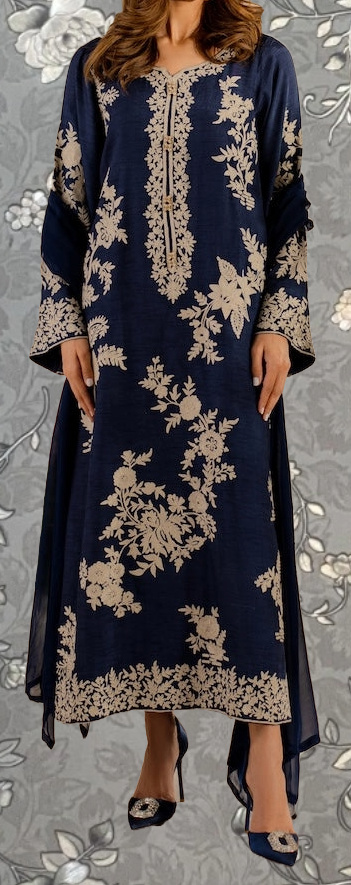 A woman wearing a long navy blue dress with gold floral embroidery, standing against a gray floral background.