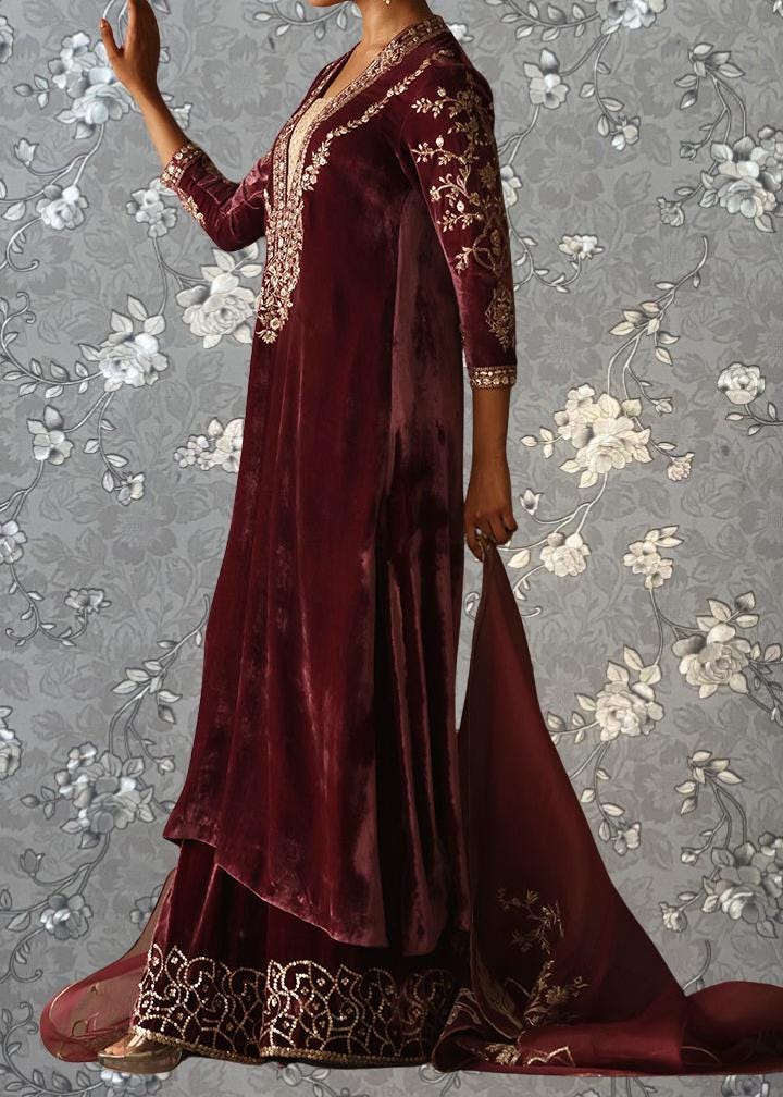 A woman wearing a long, burgundy velvet dress with intricate embroidery and a matching skirt, standing against a gray floral background.