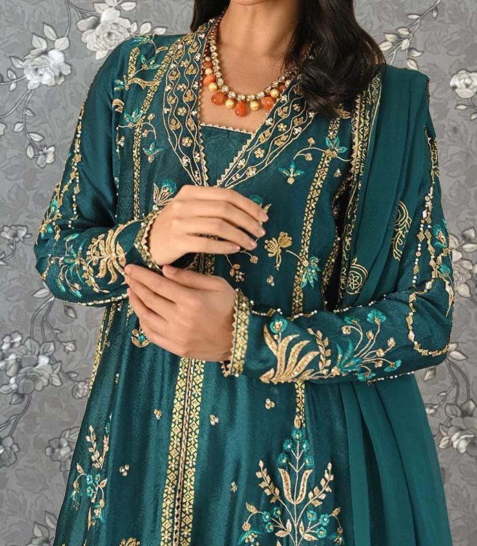A woman wearing a long, green dress with intricate gold embroidery and a matching green shawl. She is adjusting her dress and has her hands visible.