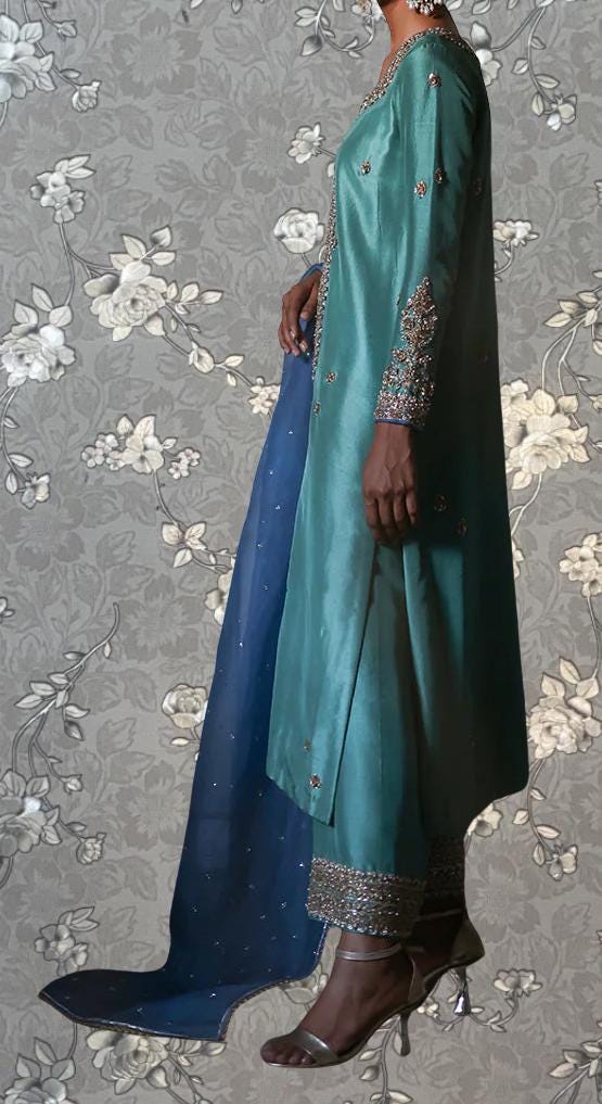 A person wearing a long, green dress with intricate embroidery and a matching blue skirt stands against a gray floral background.