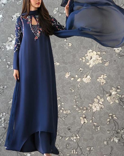 A woman wearing a long blue dress with floral embroidery poses against a gray floral background.