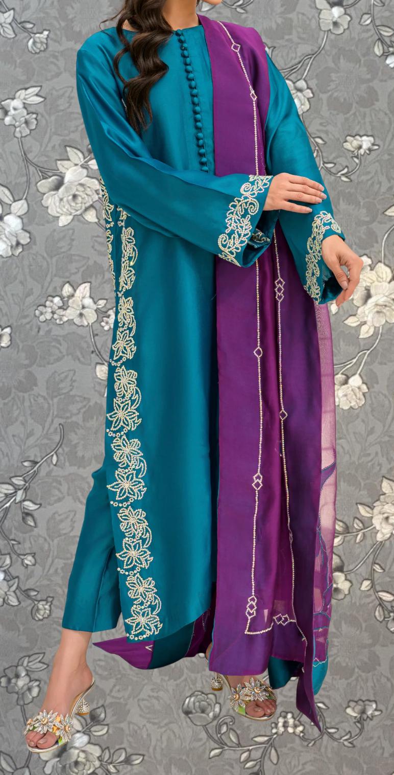 A woman wearing a long, teal-colored dress with purple accents and gold embroidery stands against a gray floral background.