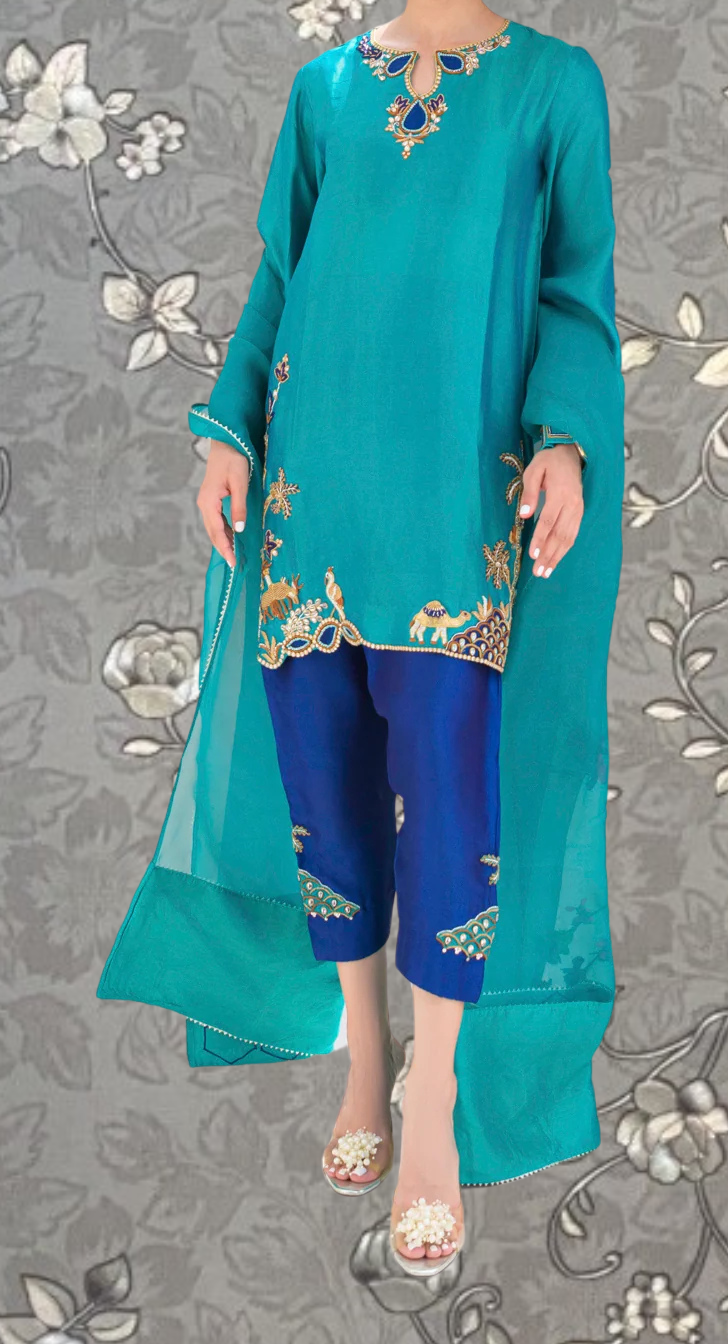 A person wearing a turquoise dress with gold embroidery and blue pants, standing against a gray floral background.