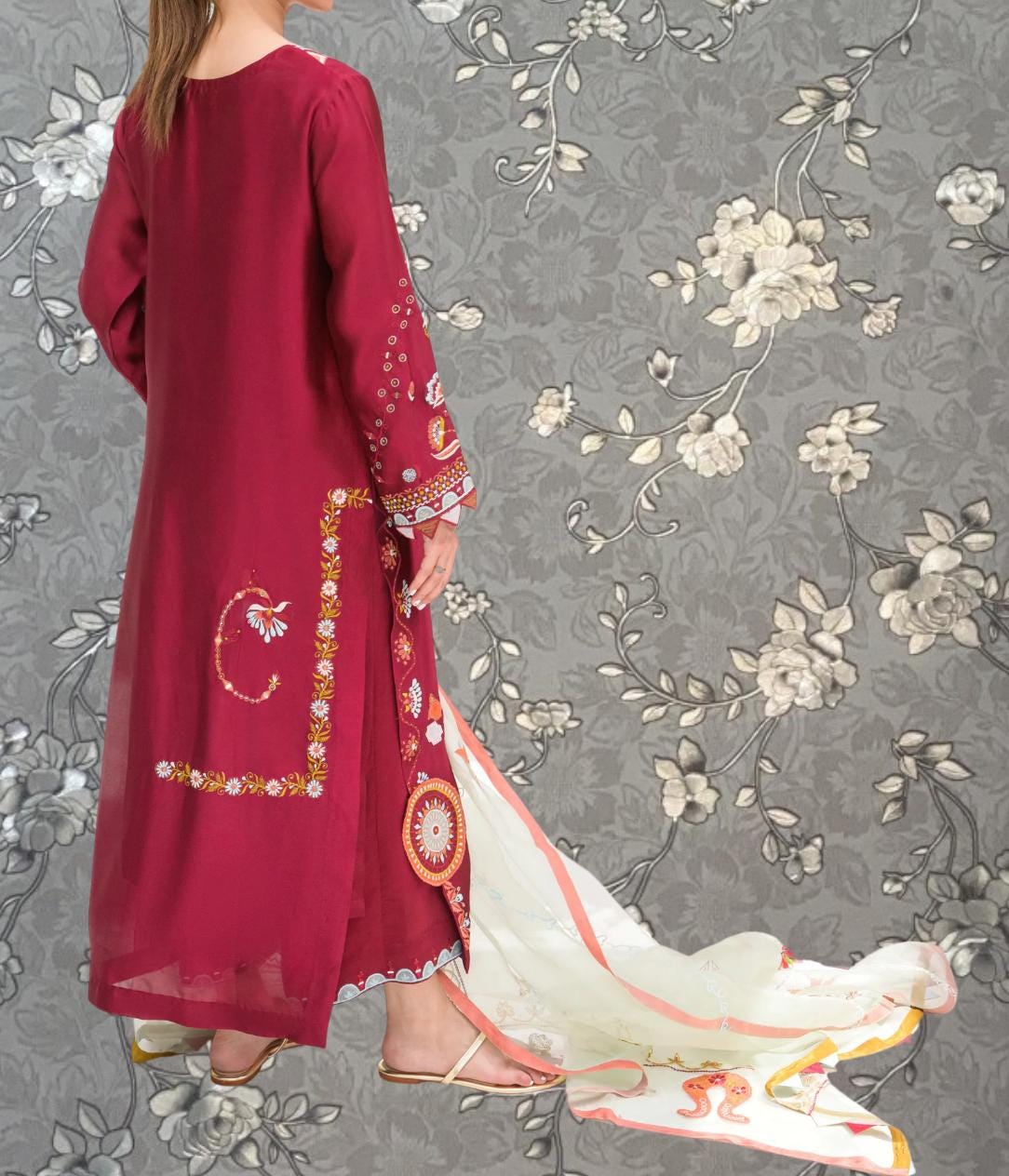 A woman wearing a long red dress with intricate floral embroidery stands against a gray floral background.