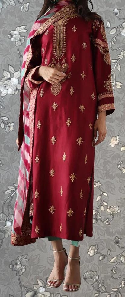 A woman wearing a red and gold embroidered dress with a matching shawl, standing against a gray floral background.