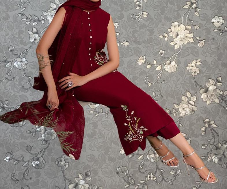 A woman wearing a red dress with floral embroidery is sitting on a chair against a gray floral background.