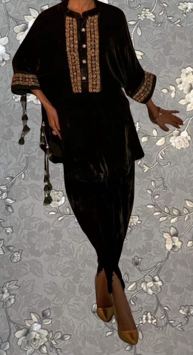 A person wearing a black velvet dress with gold embroidery and tassels, standing against a floral patterned background.