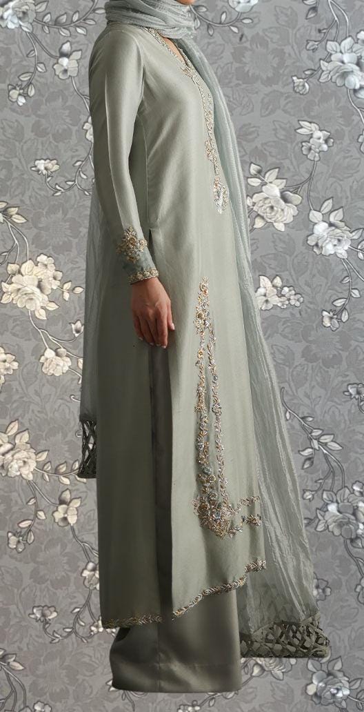A person wearing a long, gray dress with intricate embroidery and a matching gray scarf stands against a gray floral background.