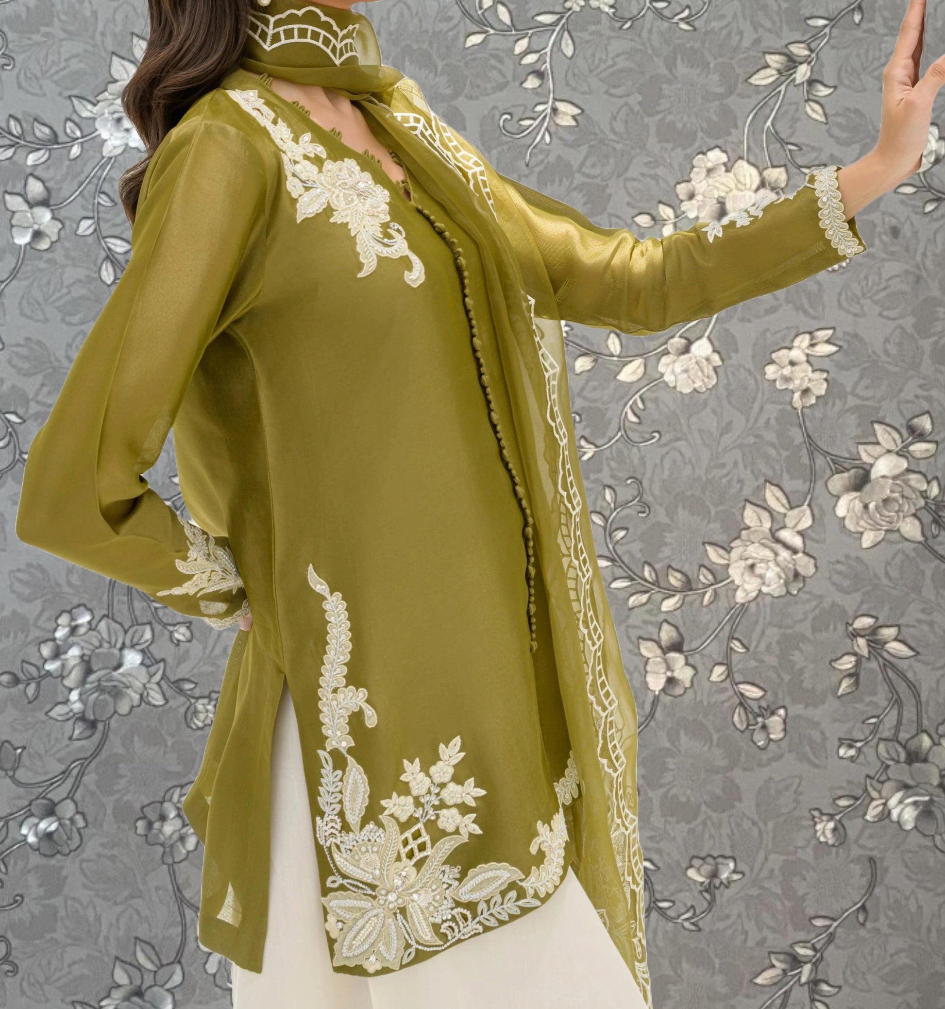 A woman wearing a green dress with white embroidered patterns poses against a gray floral background.