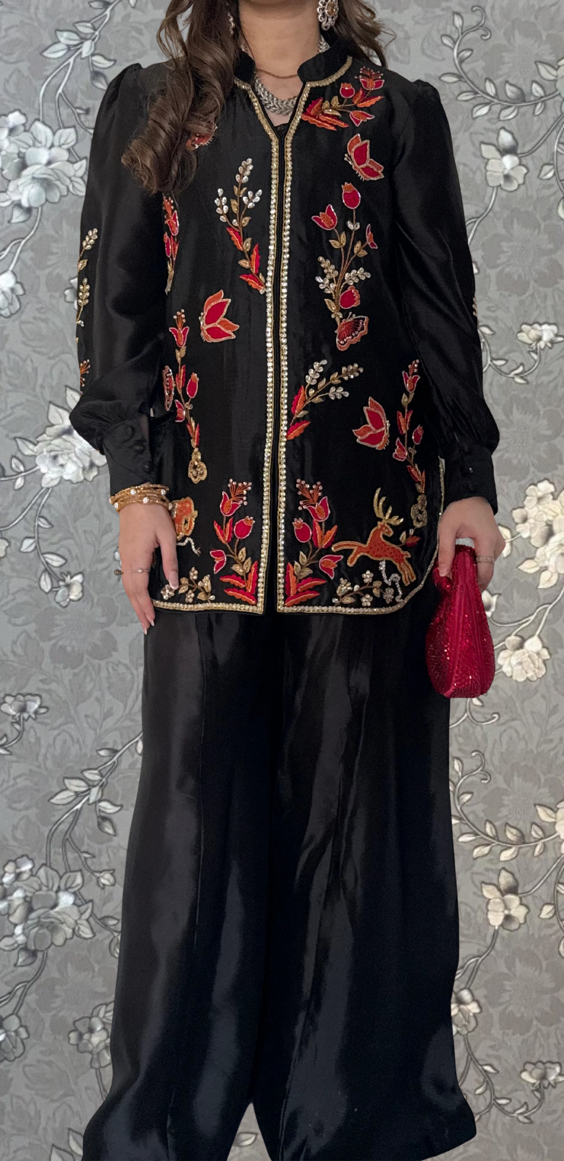 A woman wearing a black dress with red and gold embroidered patterns, standing against a floral background.