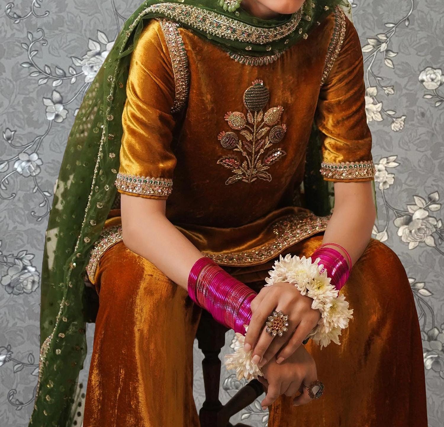 A person wearing a gold and green outfit with a floral pattern, sitting on a chair and holding a flower garland.