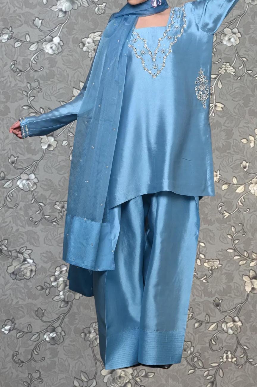 A person wearing a blue dress with a floral pattern and a matching blue scarf is standing against a gray floral background.