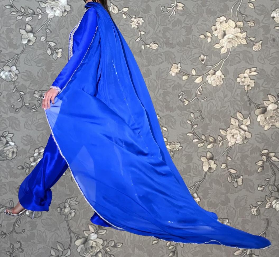 A person wearing a long, flowing blue dress with a long train walks against a gray floral background.