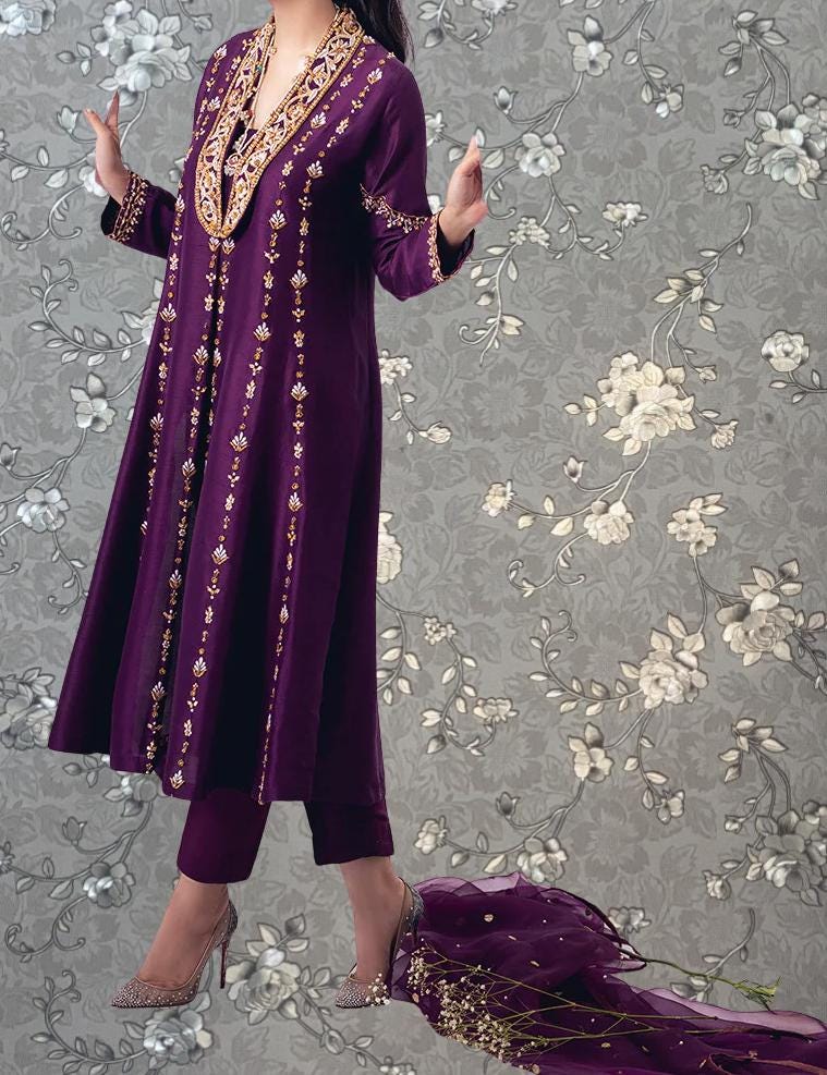 A woman wearing a long purple dress with gold embroidery stands in front of a gray floral background.