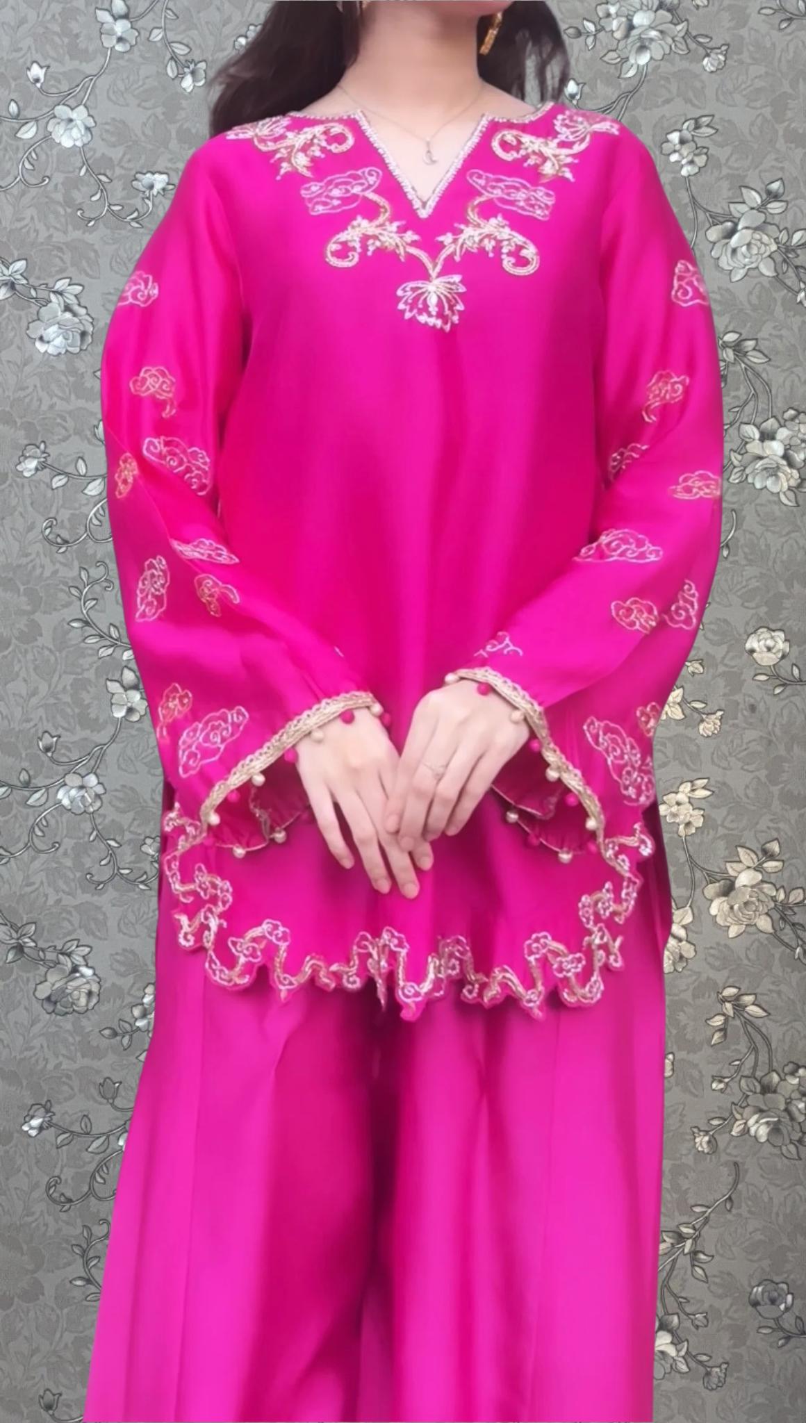 A woman wearing a bright pink dress with intricate embroidery stands against a floral background.