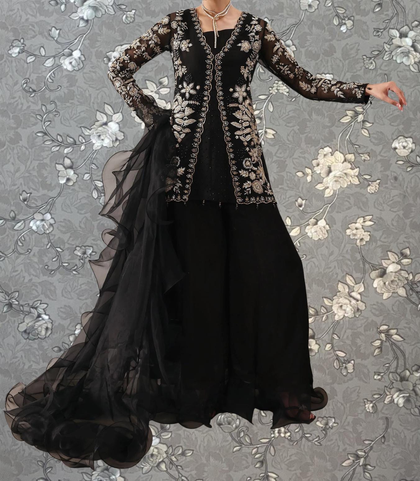 A woman wearing a long black dress with intricate embroidery and a flowing train, standing against a gray floral background.