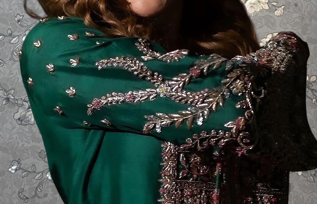 a close-up of a person wearing a green dress with intricate embroidered details. The dress has a dark green color and features a floral pattern on the sleeves and collar. The person's face is not visible in the image.
