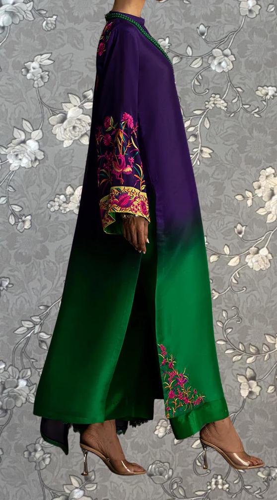 A person wearing a long, colorful dress with green and purple accents, standing against a gray floral background.