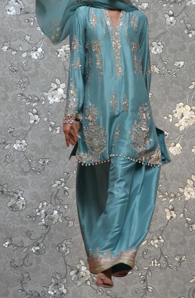 A person wearing a blue and gold embroidered dress with a floral pattern, standing against a gray and white floral background.