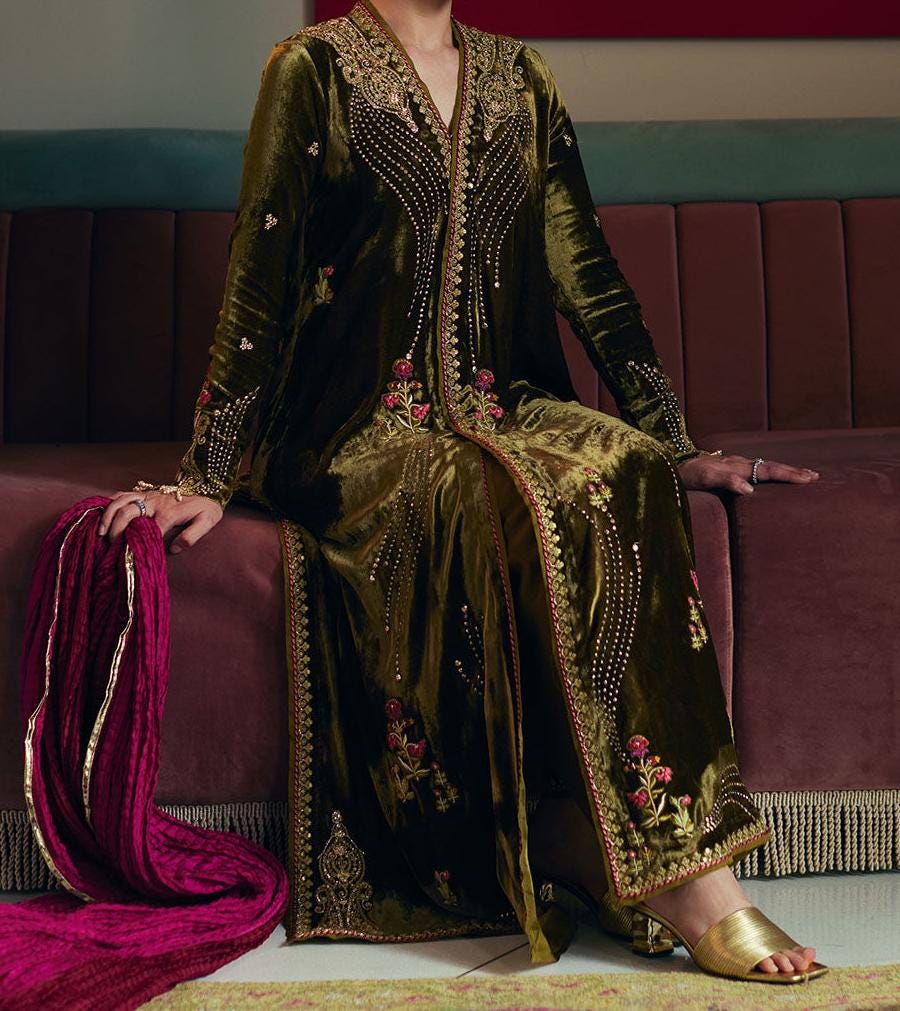 A person wearing an ornate, gold-embroidered dress is sitting on a red couch, with a pink scarf draped over their lap.