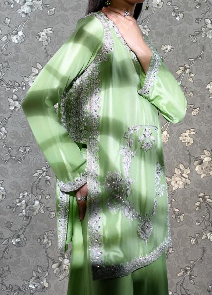 A woman wearing a green and white patterned dress with intricate embroidery stands against a gray floral background.