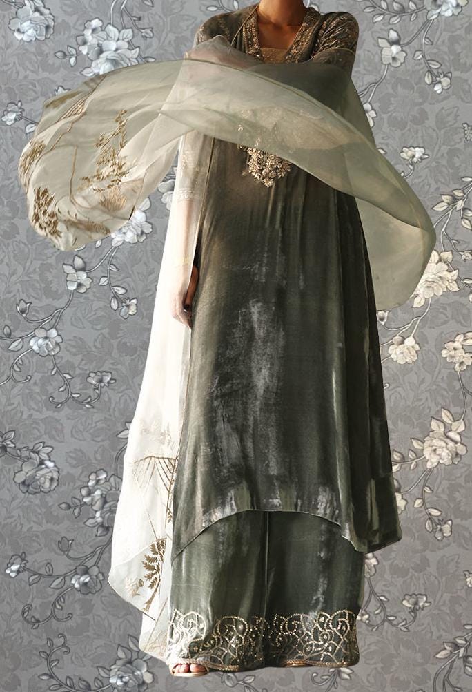 A person wearing a long, flowing gray dress with intricate gold embroidery stands against a gray floral background.