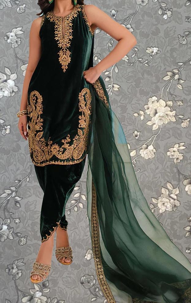 A woman wearing a green velvet dress with gold embroidery and a matching green shawl, standing against a gray floral background.