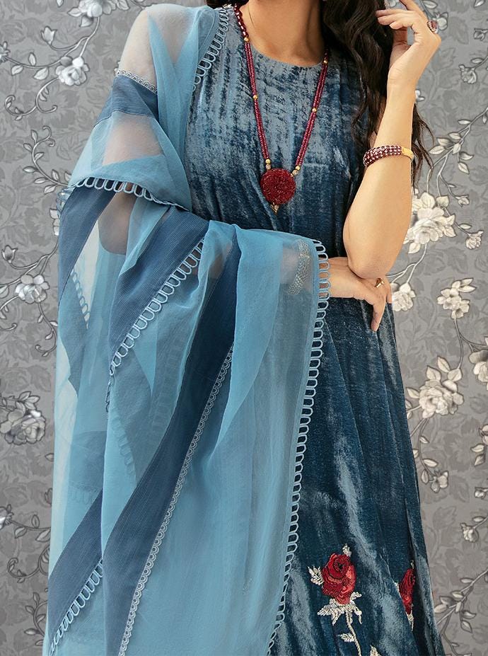 A woman wearing a blue dress with red embroidered details and a matching blue shawl. She is also wearing a necklace with red beads.