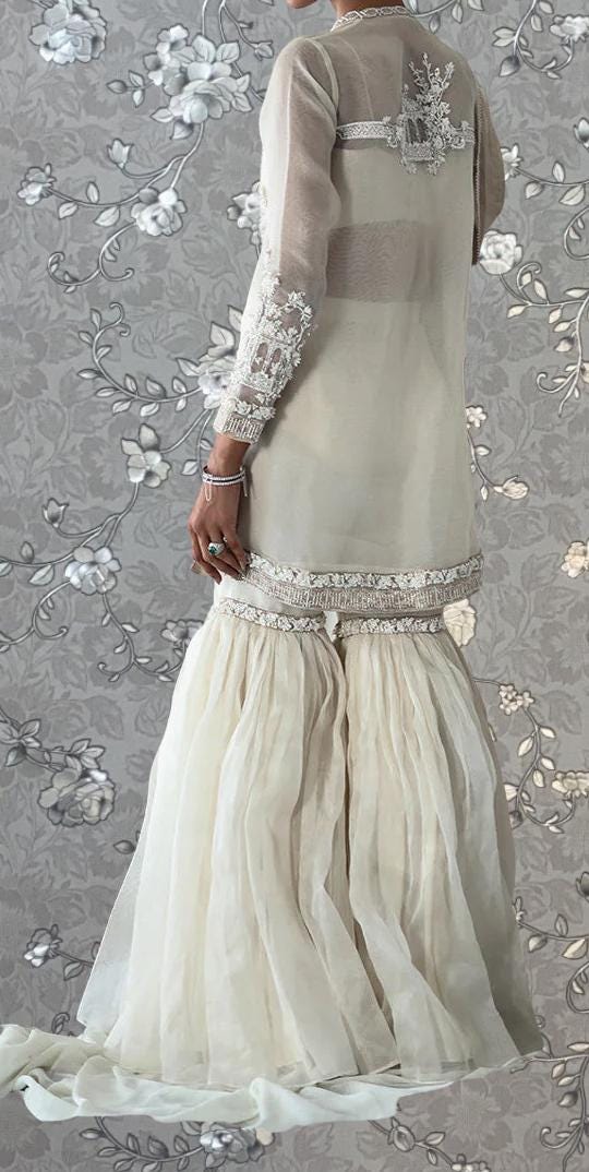 A person wearing a long, white dress with lace sleeves and a high skirt, standing against a gray floral background.