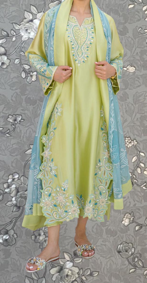 A woman wearing a long, yellow dress with intricate embroidery and a matching blue shawl. She is standing against a gray floral background.