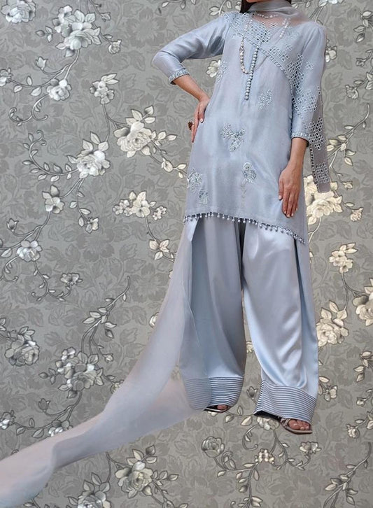 A woman wearing a light blue dress with intricate embroidery and a flowing train, standing against a gray floral background.