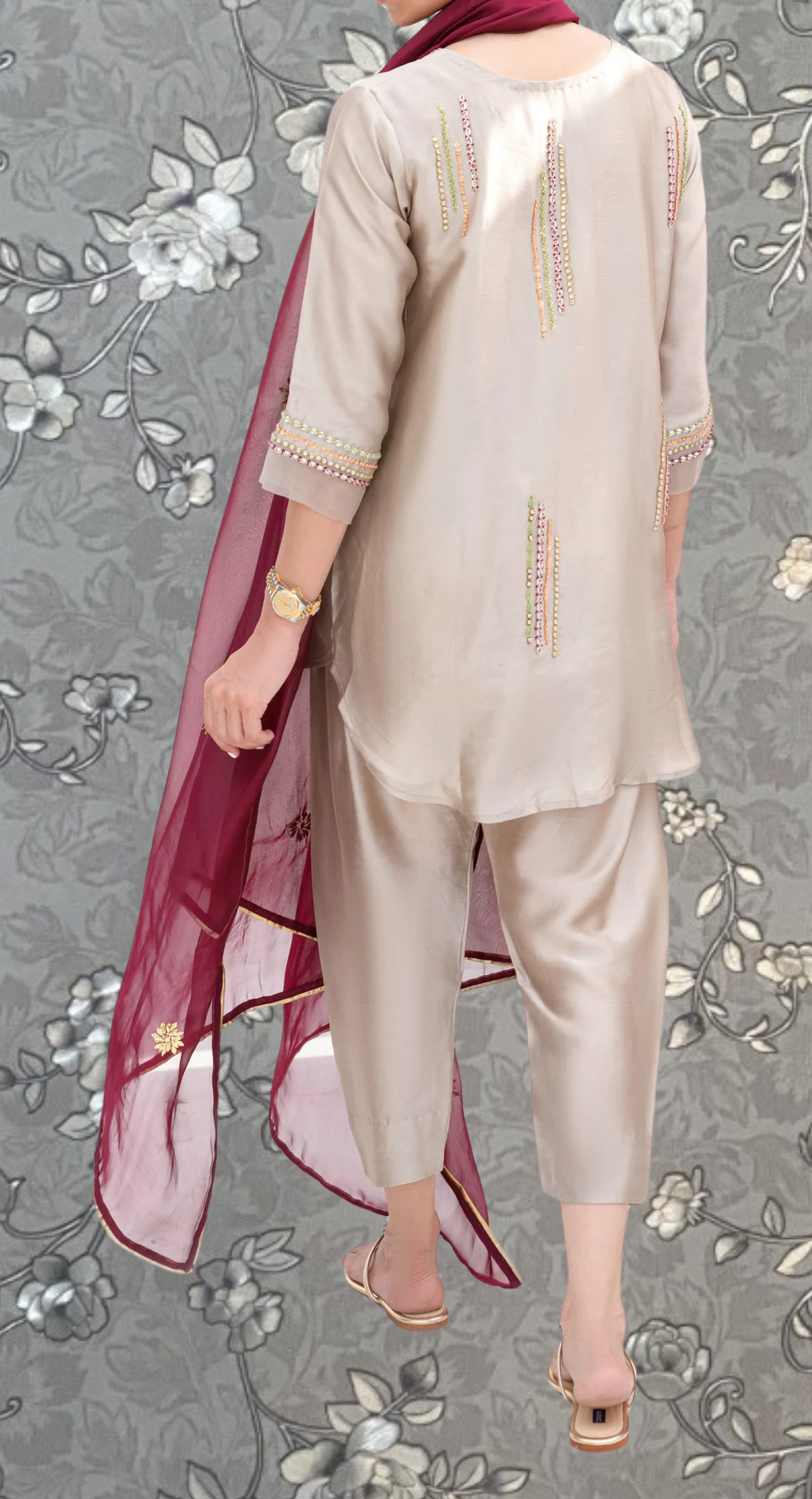 A person wearing a beige outfit with a red shawl and gold accessories, standing against a gray floral background.