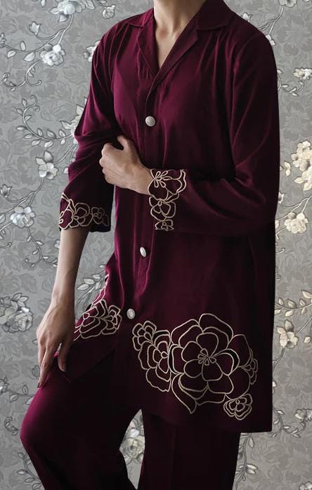 A person wearing a burgundy outfit with gold floral embroidery stands against a gray floral background.