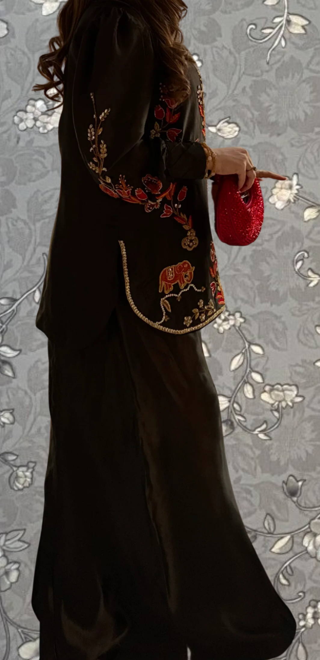 A woman wearing a black dress with red floral embroidery and holding a red purse stands against a gray floral background.
