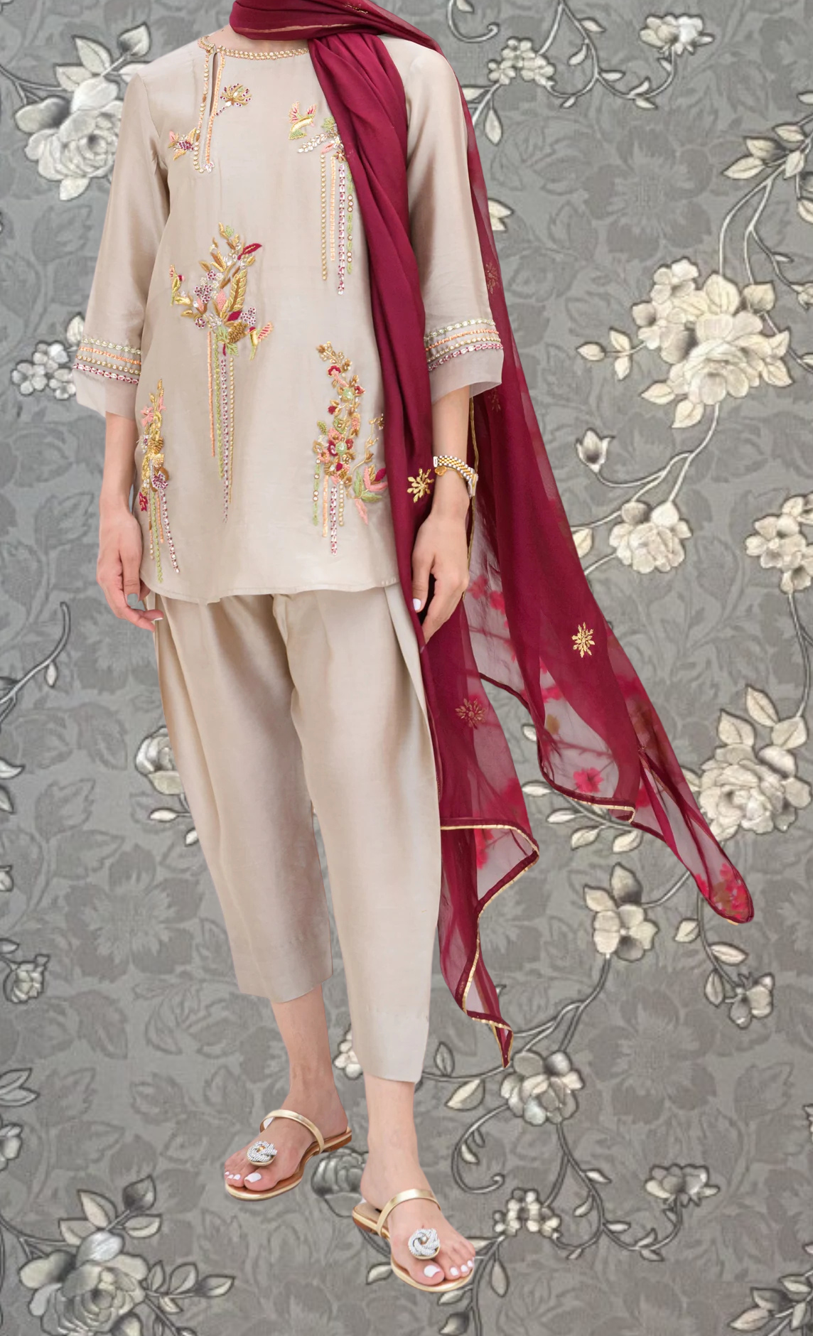 A person wearing a beige outfit with gold embroidery and a red shawl stands against a gray floral background.