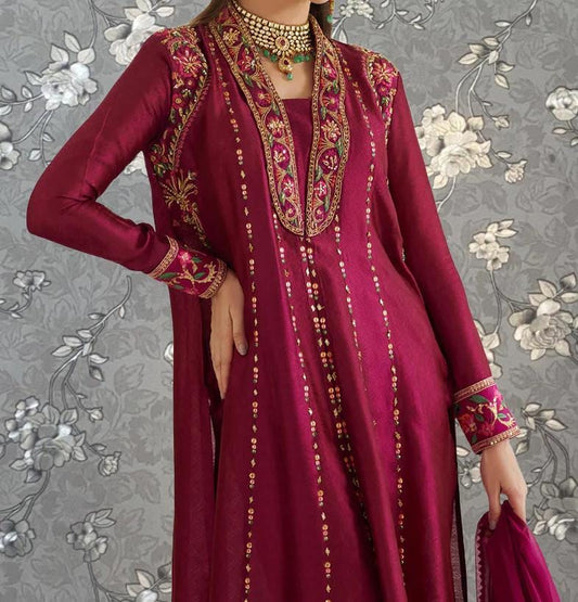 A woman wearing a long, burgundy dress with intricate embroidery and gold accents, posing against a gray floral background.