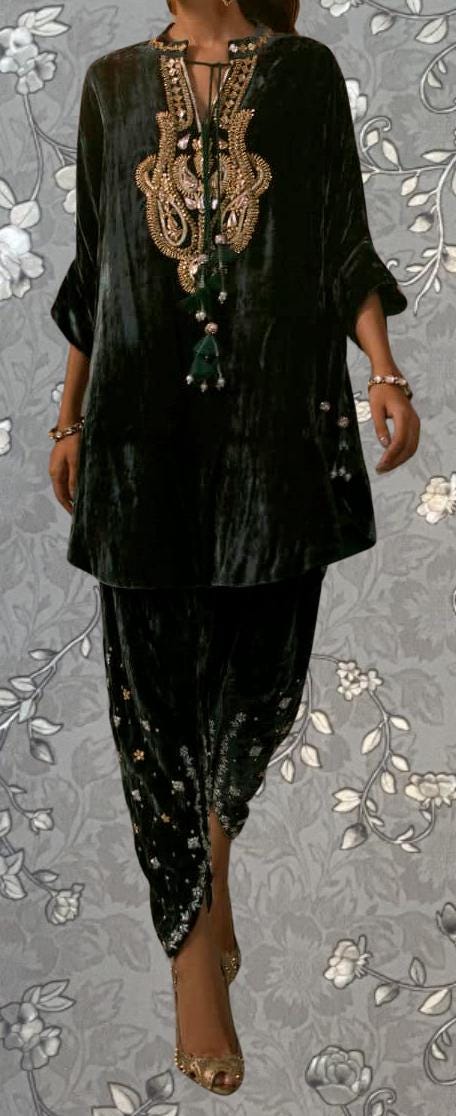 A person wearing a black velvet dress with intricate gold embroidery and tassels, standing against a gray floral background.