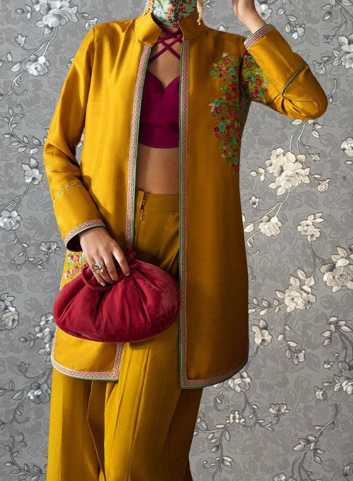 A person wearing a yellow jacket with floral embroidery and a red purse is standing against a gray floral background.