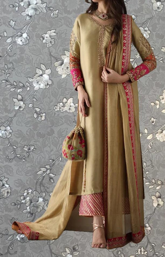A woman wearing a gold and pink traditional Indian outfit, including a long skirt and a shawl, stands against a gray floral background. She is holding a small purse in her hand.