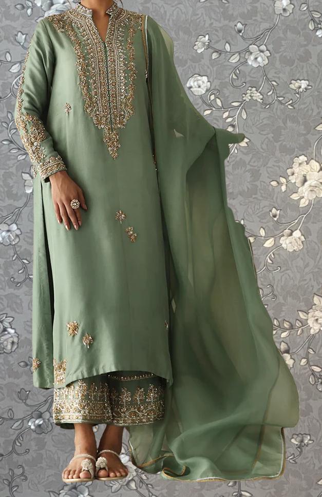 A person wearing a green and gold embroidered dress stands against a gray floral background.