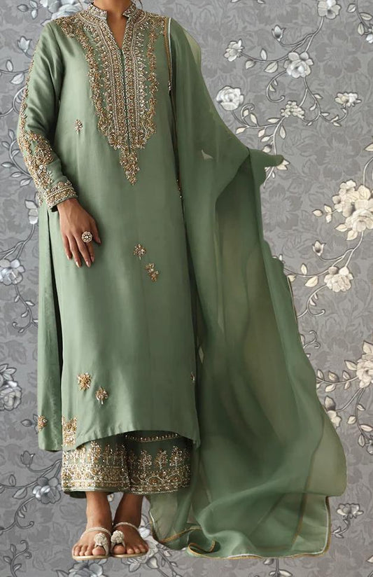 A person wearing a green and gold embroidered dress stands against a gray floral background.