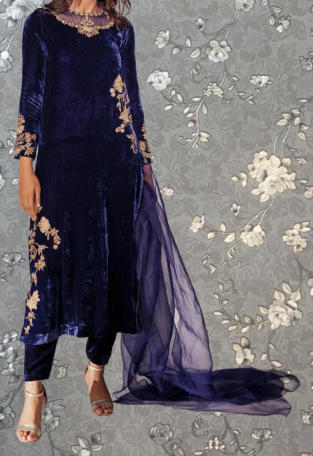 A woman wearing a long, blue velvet dress with gold embroidery stands against a gray floral background.